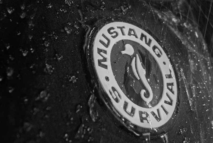 Close-up of a round, waterproof Mustang Survival seahorse logo patch on wet black technical fabric covered in water droplets.