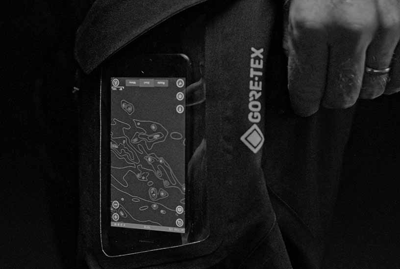 Black and white close-up of a smartphone displaying a topographic map inside a clear sleeve on a Gore-Tex jacket pocket.