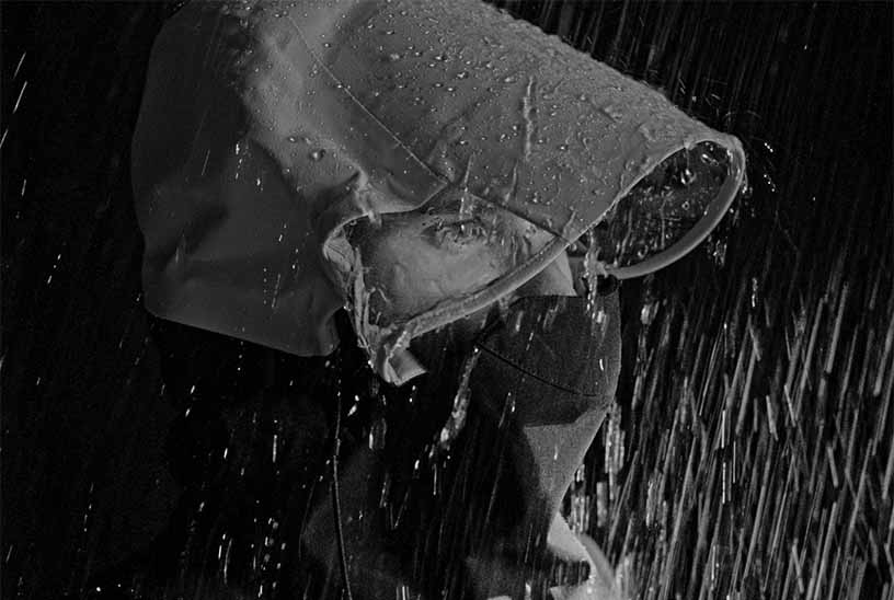 Black & white close-up of a person wearing a protective hood with side windows & collar protection under heavy simulated rain.